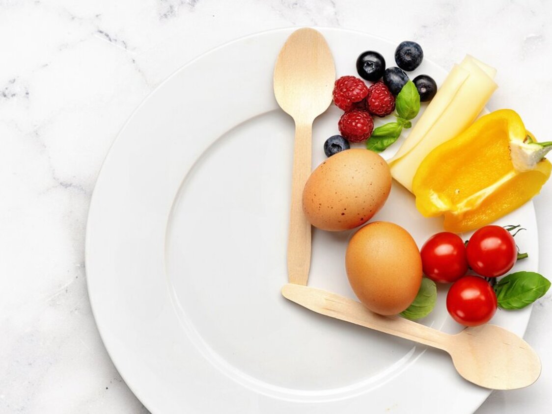 Ein Teller mit zwei Holzlöffeln, was die Optik einer Uhr bildet. Zwischen beiden Löffeln Gemüse, Obst und Eier.
