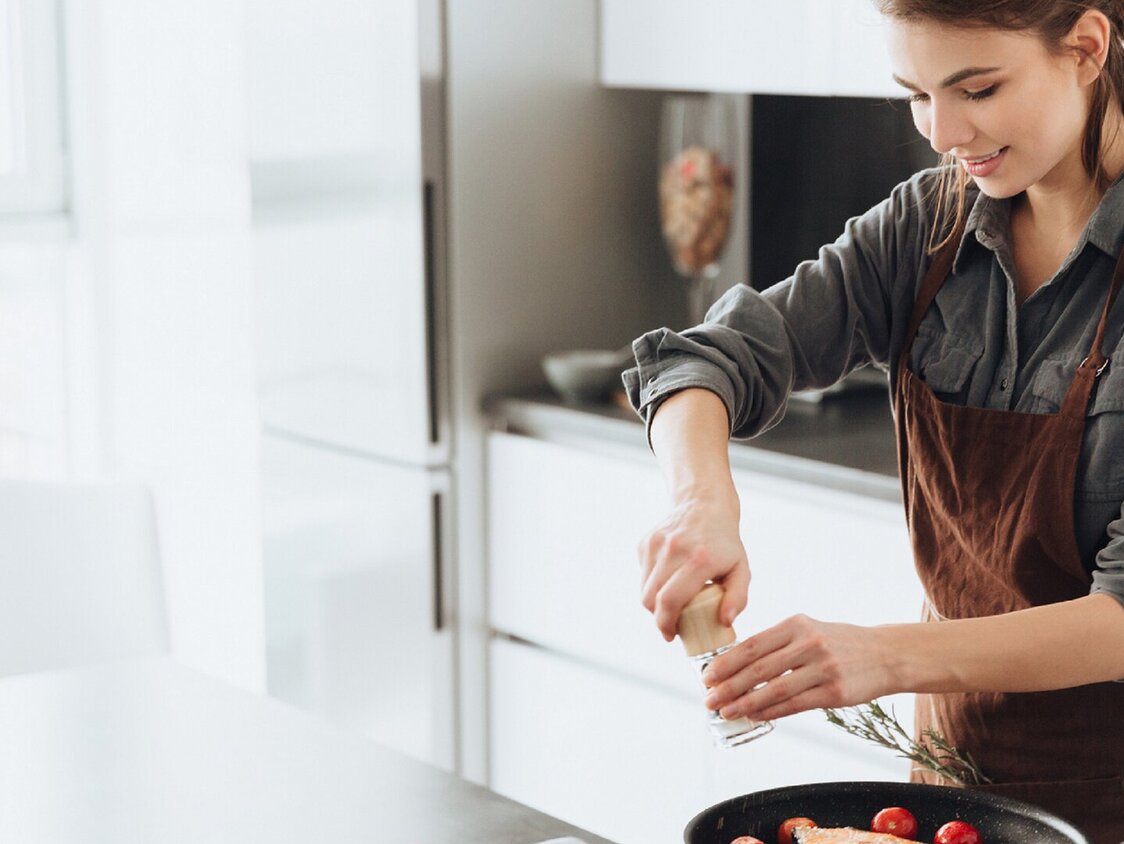 Eine Frau steht in der Küche am Herd und würzt Fisch in einer Pfanne.