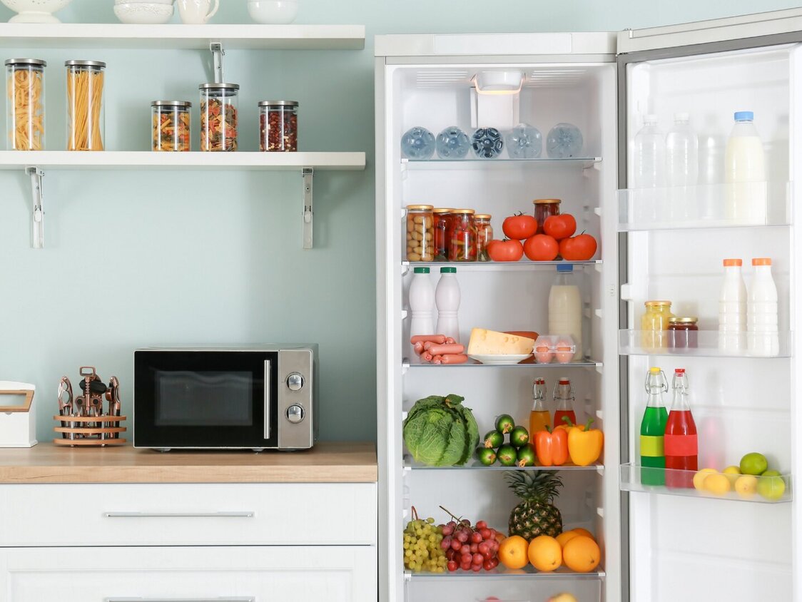 Eine Küchenzeile mit einem geöffneten Kühlschrank. Er ist voller Obst, Gemüse und Milch. Auf der Küchenzeile Kochutensilien und eine Mikrowelle.