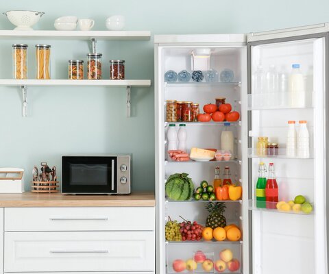 Eine Küchenzeile mit einem geöffneten Kühlschrank. Er ist voller Obst, Gemüse und Milch. Auf der Küchenzeile Kochutensilien und eine Mikrowelle.