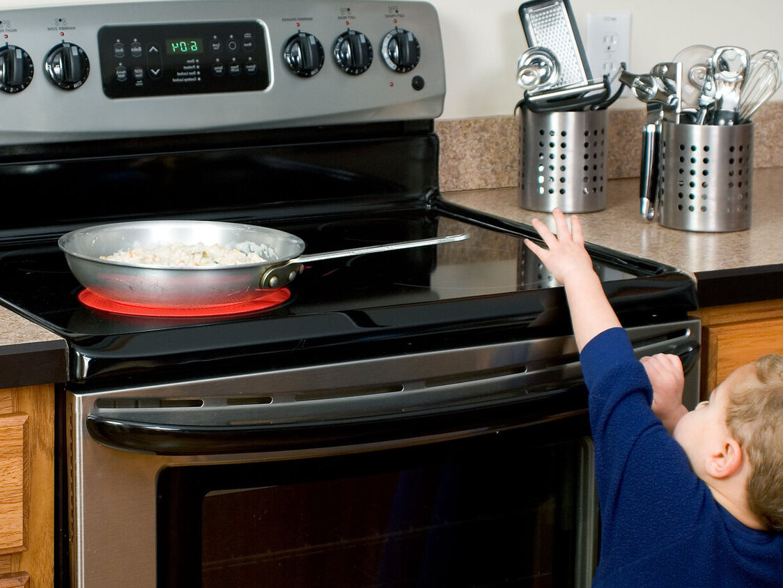 Ein kleines Kind greift hoch zu einer Pfanne, die auf einer heißen Herdplatte steht.