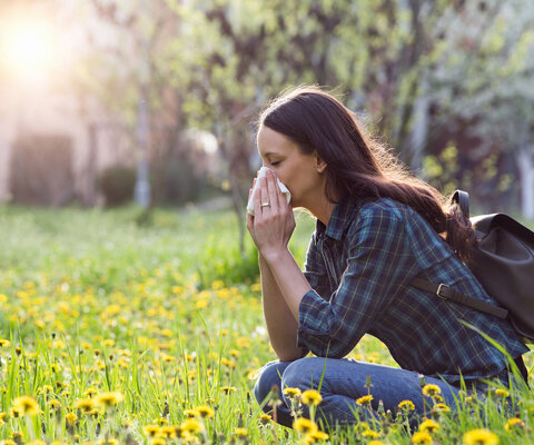 Eine Frau auf einer Blumenwiese hockend. Sie putzt ihre Nase. Bäume und die Sonne im Hintergrund.