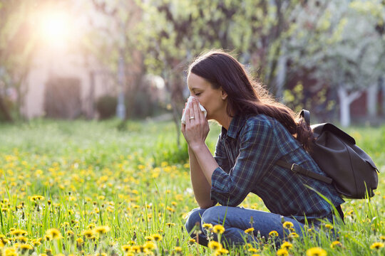 1067954744 Eine Frau auf einer Blumenwiese hockend. Sie putzt ihre Nase. Bäume und die Sonne im Hintergrund.