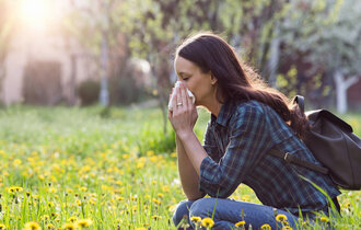 Eine Frau auf einer Blumenwiese hocken. Sie putzt ihre Nase. Bäume und die Sonne im Hintergrund.