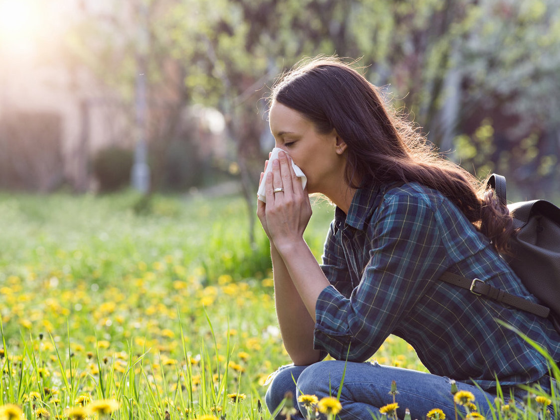 Eine Frau auf einer Blumenwiese hockend. Sie putzt ihre Nase. Bäume und die Sonne im Hintergrund.