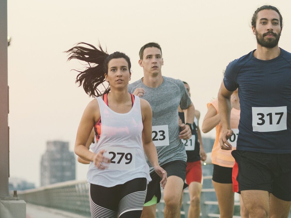 Mehrere Personen mit Startnummern auf ihren Shirts laufen auf einer Brücke.