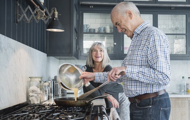 Ein alter Mann macht essen, während eine alte Frau zuguckt.