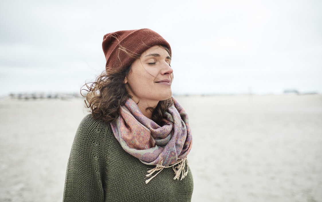 Portrait eine lächelnden Frau mit Mütze und Schal am Strand.