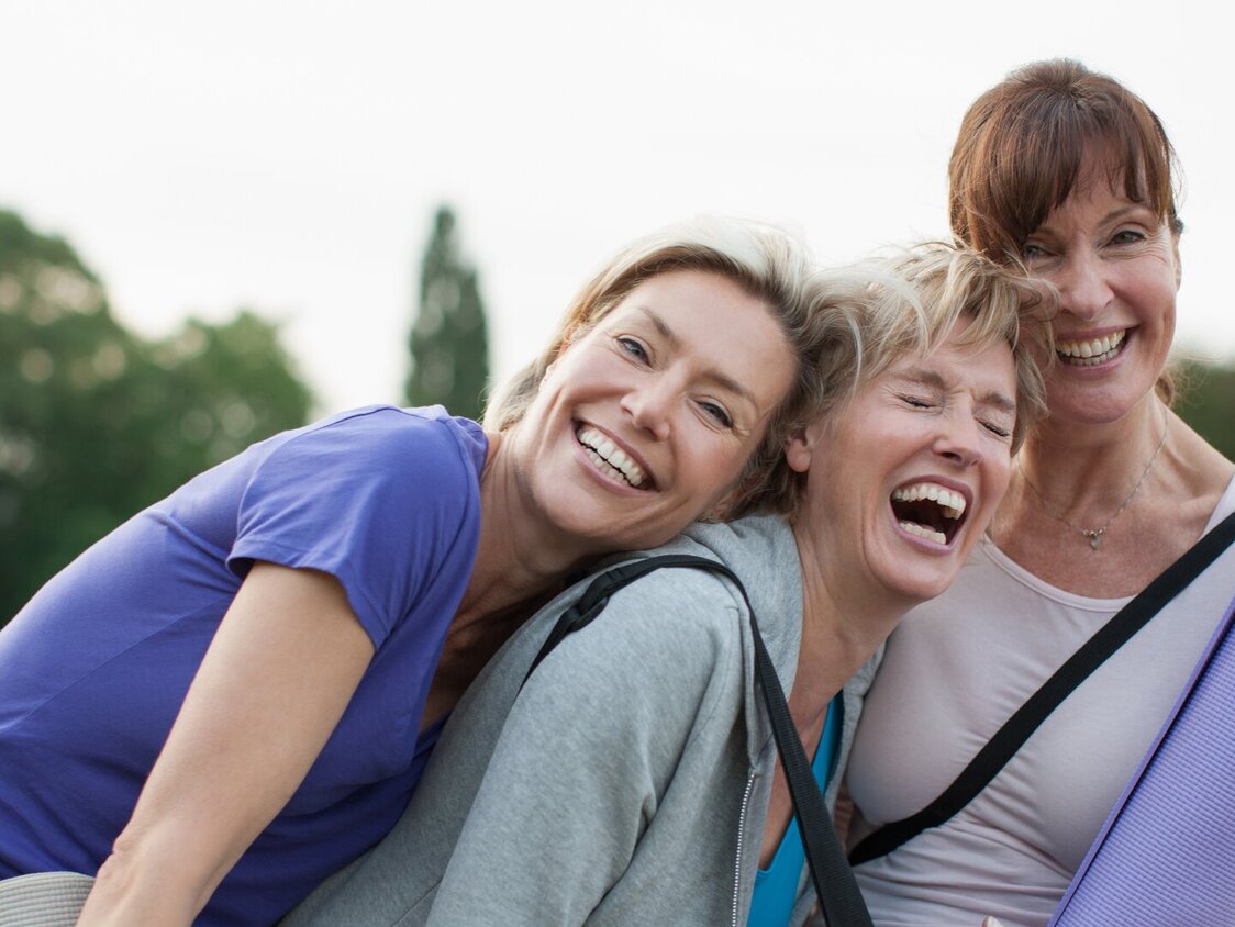 Drei Frauen mit Yogamatten in der Hand, lehnen gegeneinander und lachen.
