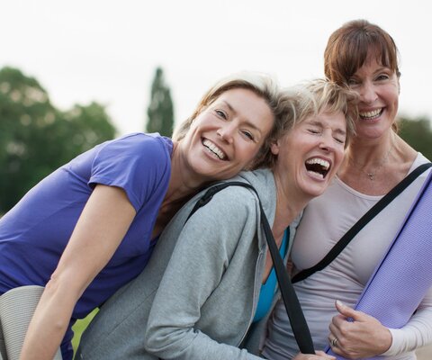 Drei Frauen mit Yogamatten in der Hand, lehnen gegeneinander und lachen.