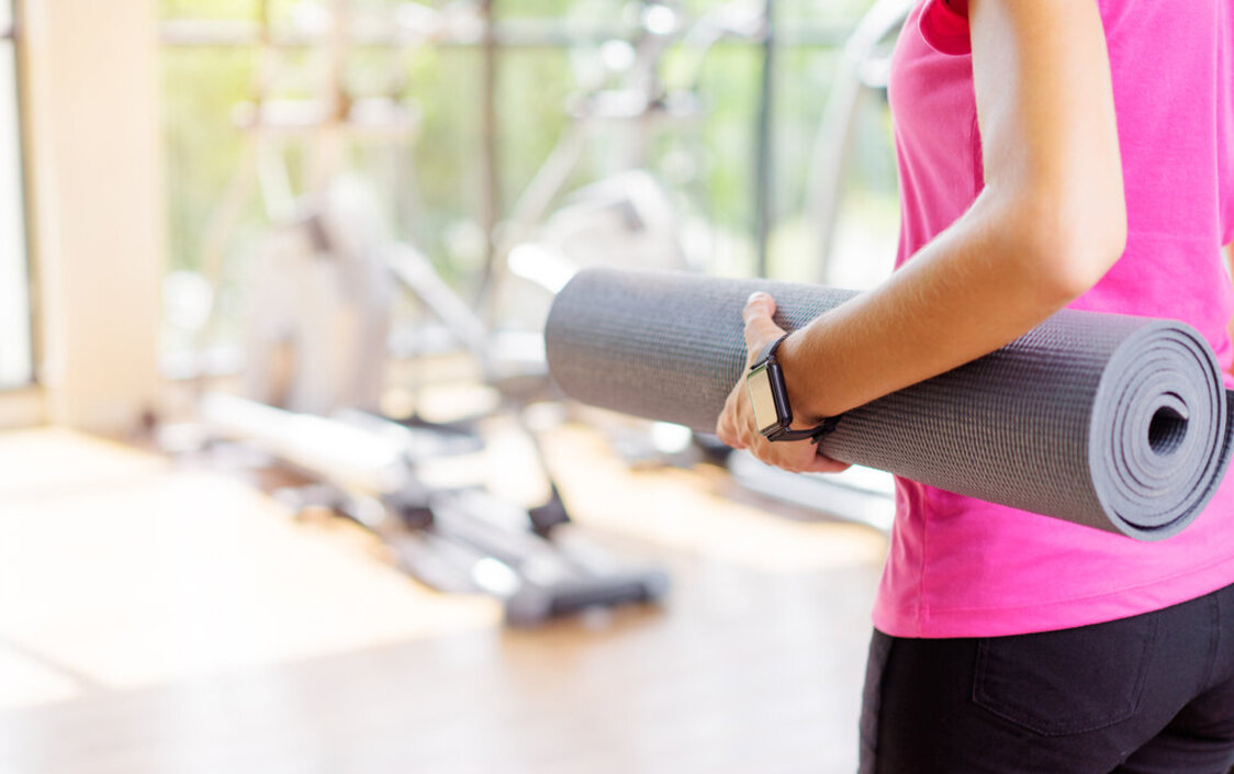 Eine Frau mit pinkem Sport-Top hält eine Yogamatte unter dem Arm und steht in einem Studio. Man sieht nur ihren Rücken.