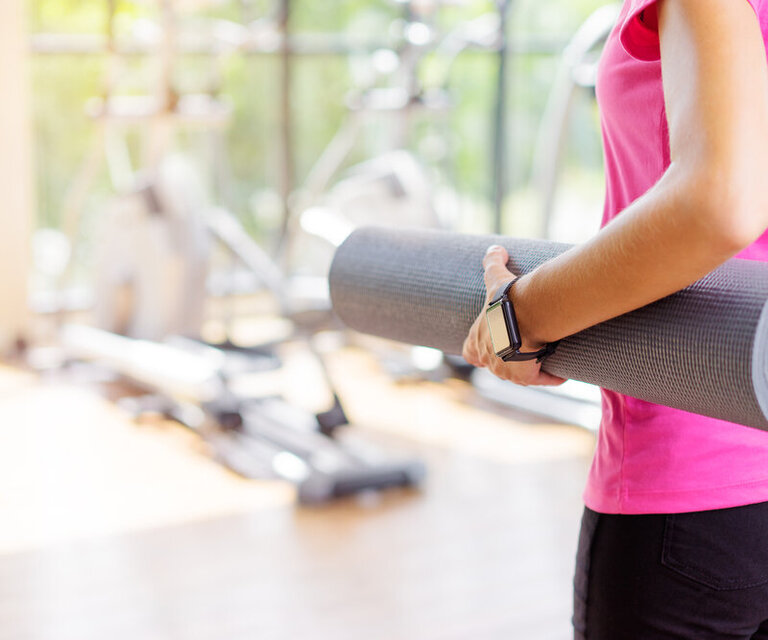 Eine Frau mit pinkem Sport-Top hält eine Yogamatte unter dem Arm und steht in einem Studio. Man sieht nur ihren Rücken.