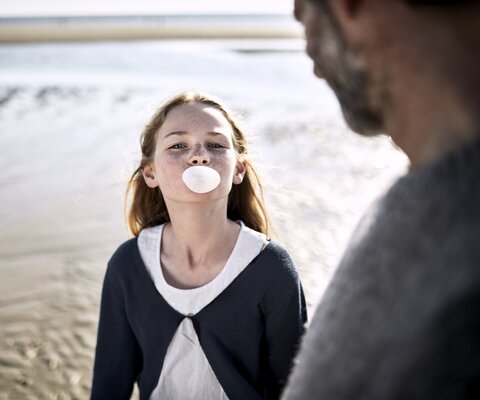 Ein Mädchen steht am Strand und macht eine Kaugummiblase. Ein Mann steht daneben.