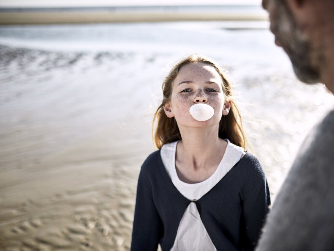Ein Mädchen steht am Strand und macht eine Kaugummiblase. Ein Mann steht daneben.