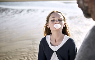 Ein Mädchen steht am Strand und macht eine Kaugummiblase. Ein Mann steht daneben.