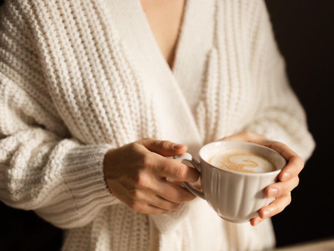 Eine Frau in einem beigen Strick-Cardigan hält eine Tasse Cappuccino.
