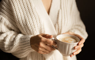 Eine Frau in einem beigen Strick-Cardigan hält eine Tasse Cappuccino.