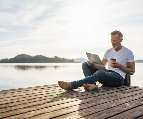 Ein Mann sitzt auf einem Steg am Wasser und liest Zeitung. Er hat einen Kaffee To-Go dabei.