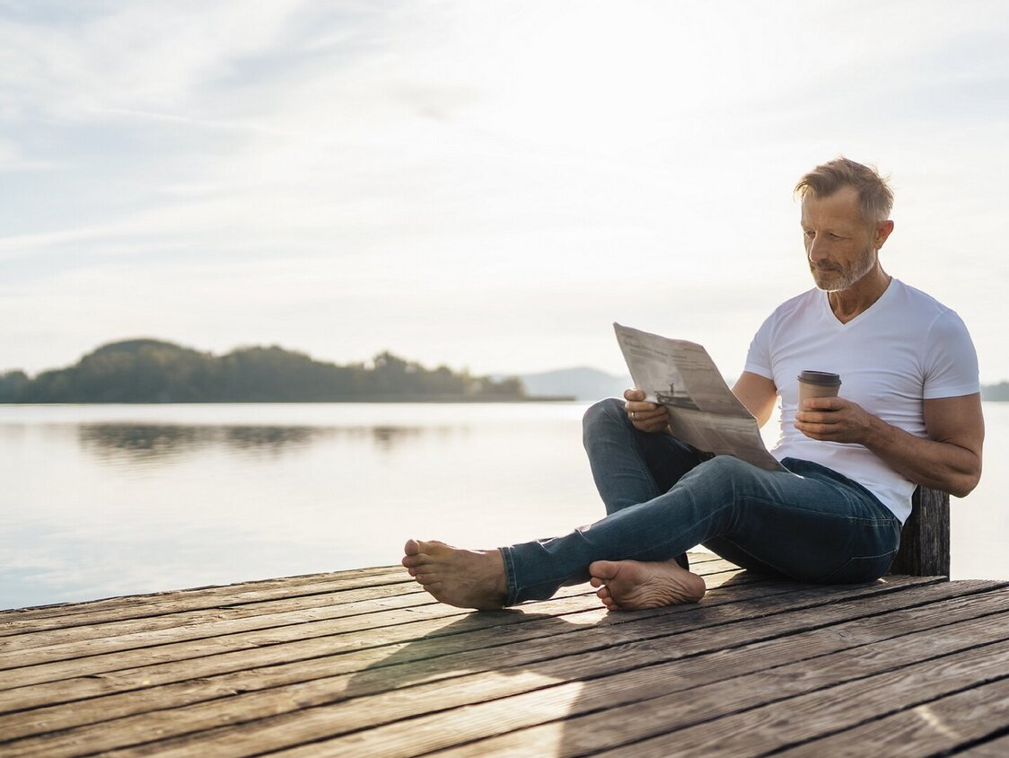 Ein Mann sitzt auf einem Steg am Wasser und liest Zeitung. Er hat einen Kaffee To-Go dabei.