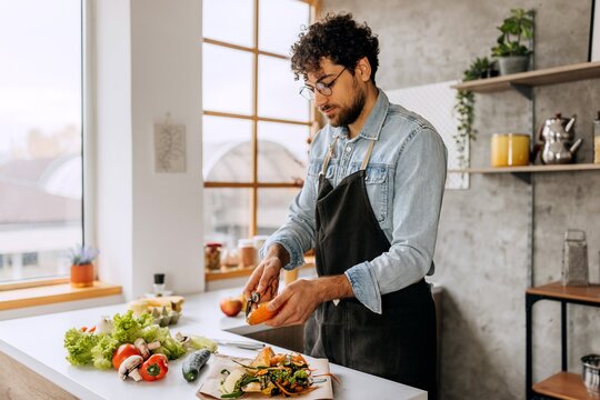  Ein Mann steht in der Küche und schält eine Möhre. Auf der Arbeitsplatte liegen eine Gurke, Paprika, Pilze und Salat.