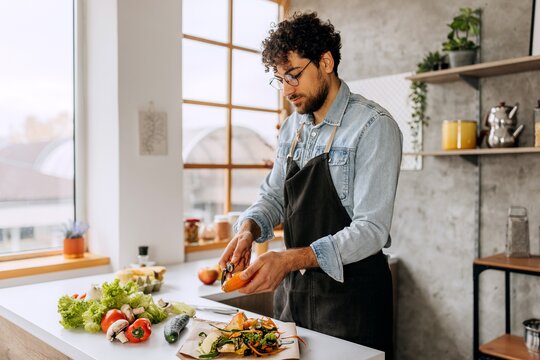 Ein Mann steht in der Küche und schält eine Möhre. Auf der Arbeitsplatte liegen eine Gurke, Paprika, Pilze und Salat.