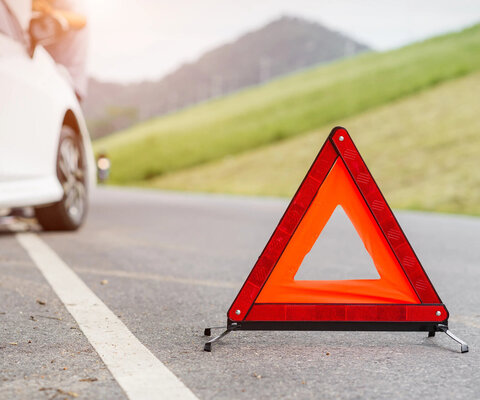 Ein Warndreieck steht auf einer Straße. Im Hintergrund ist ein weißes Auto am Straßenrand zu erkennen.