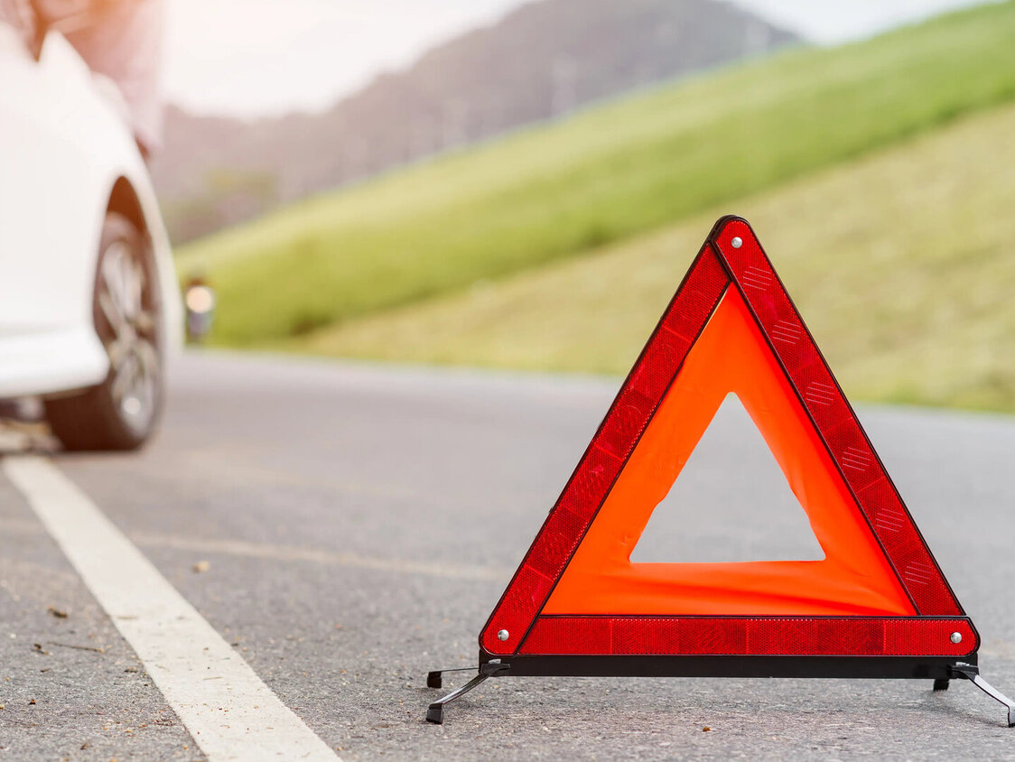Ein Warndreieck steht auf einer Straße. Im Hintergrund ist ein weißes Auto am Straßenrand zu erkennen.