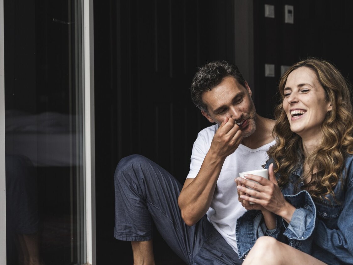 Ein Mann und eine Frau sitzend lachend an der Terasse. Sie trinkt Kaffee und er hat einen Löffel am Mund.
