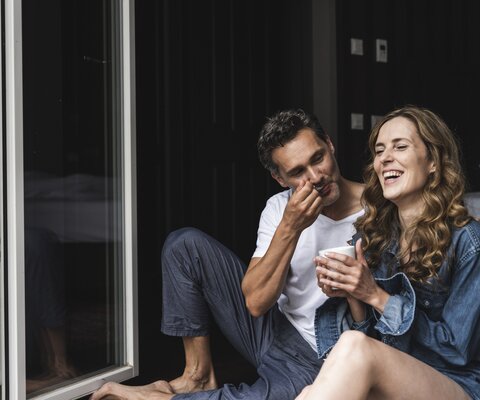 Ein Mann und eine Frau sitzend lachend an der Terasse. Sie trinkt Kaffee und er hat einen Löffel am Mund.