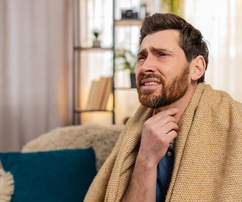 Mann sitzt mit Decke auf dem Sofa und fasst sich mit schmerzverzehrtem Gesicht an den Hals.