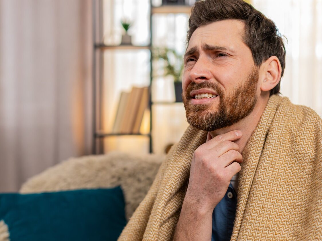 Mann sitzt mit Decke auf dem Sofa und fasst sich mit schmerzverzehrtem Gesicht an den Hals.