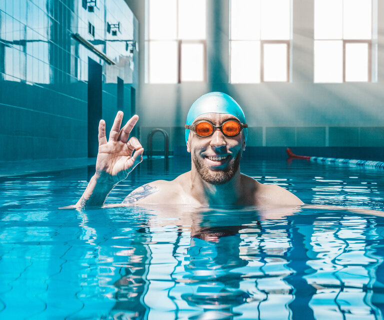 Ein Mann in einem Schwimmbecken mit Taucherbrille und Badekappe, der lächelnd das Taucherzeichen für "alles in Ordnung" anzeigt.