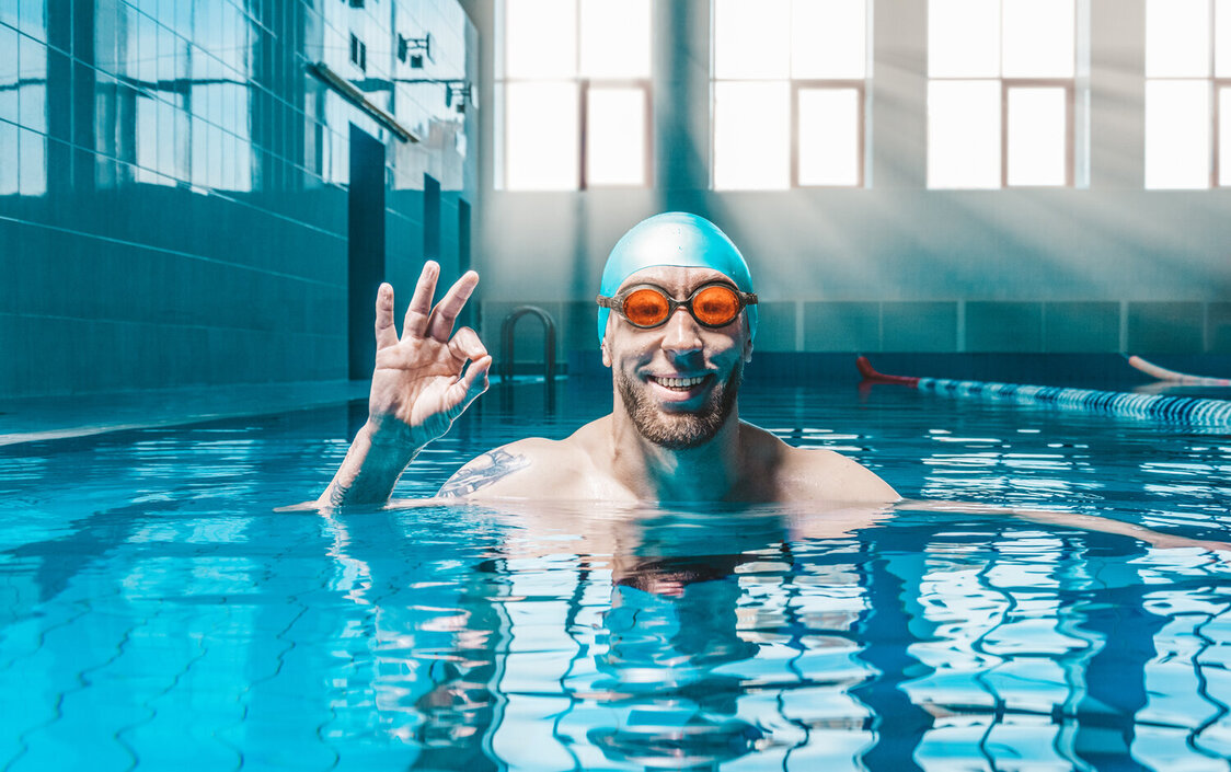 Ein Mann in einem Schwimmbecken mit Taucherbrille und Badekappe, der lächelnd das Taucherzeichen für "alles in Ordnung" anzeigt.