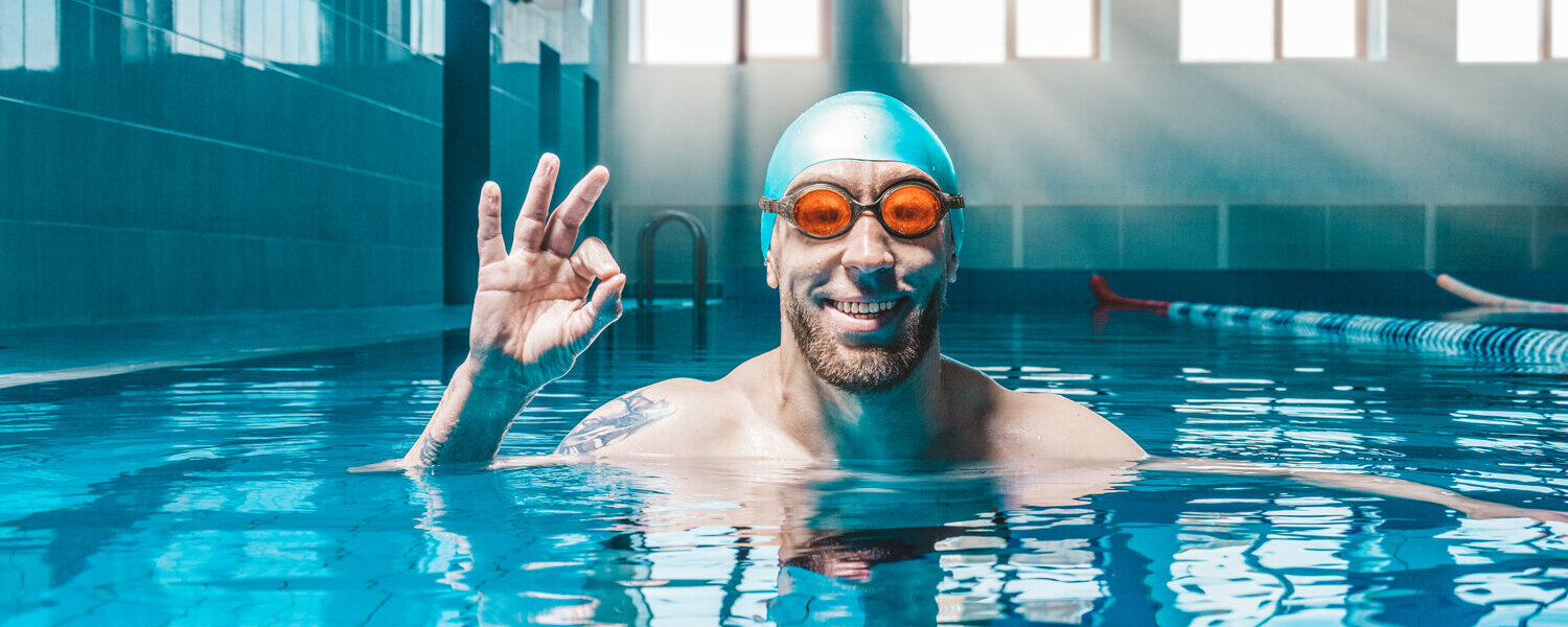 Ein Mann in einem Schwimmbecken mit Taucherbrille und Badekappe, der lächelnd das Taucherzeichen für "alles in Ordnung" anzeigt.