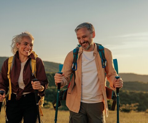 Das Bild zeigt zwei ältere Personen, die bei Sonnenuntergang mit Walking Stöcken wandern gehen.