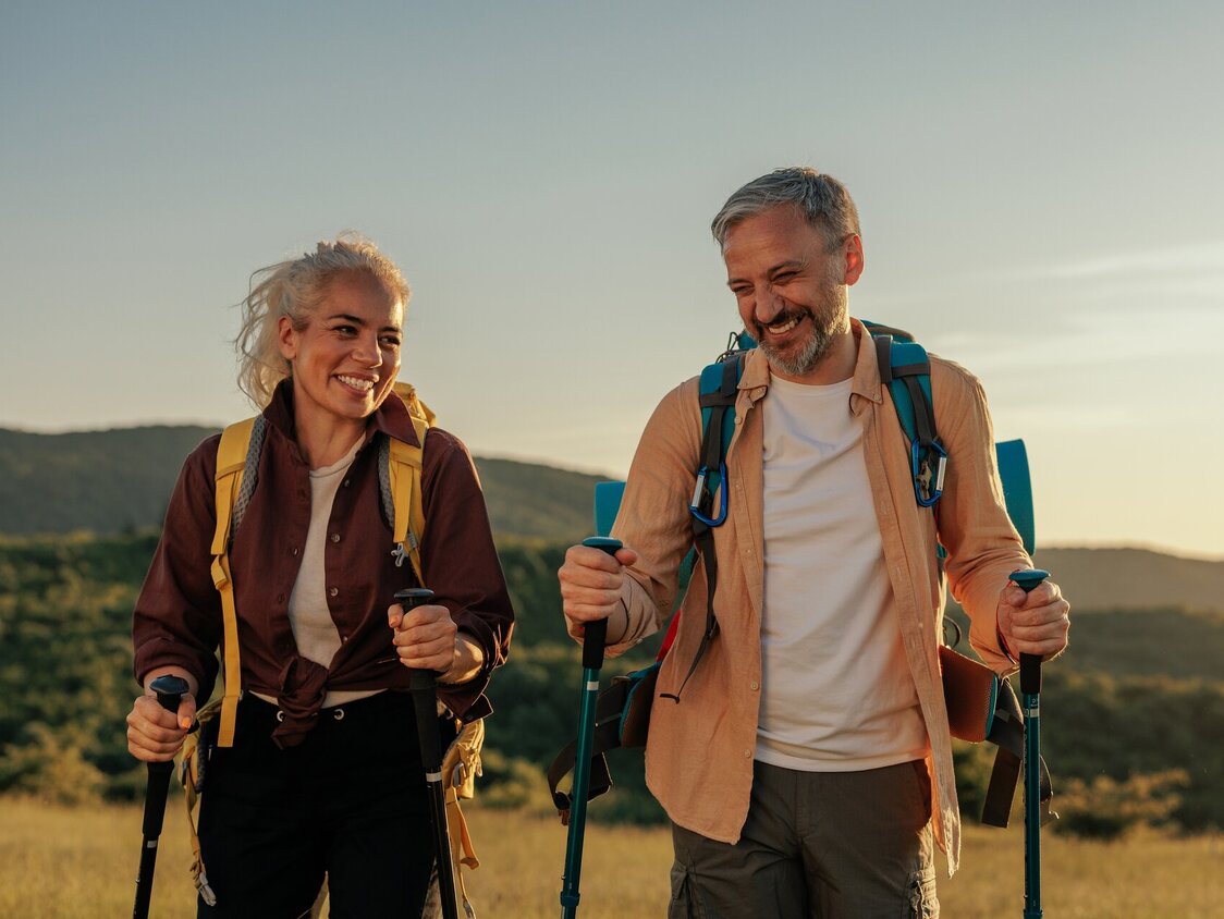 Das Bild zeigt zwei ältere Personen, die bei Sonnenuntergang mit Walking Stöcken wandern gehen.