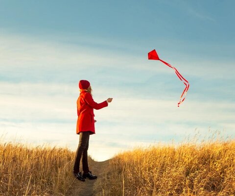 Eine Frau in einer roten Jacke lässt in einem herbstlichen Feld einen Drachen steigen.