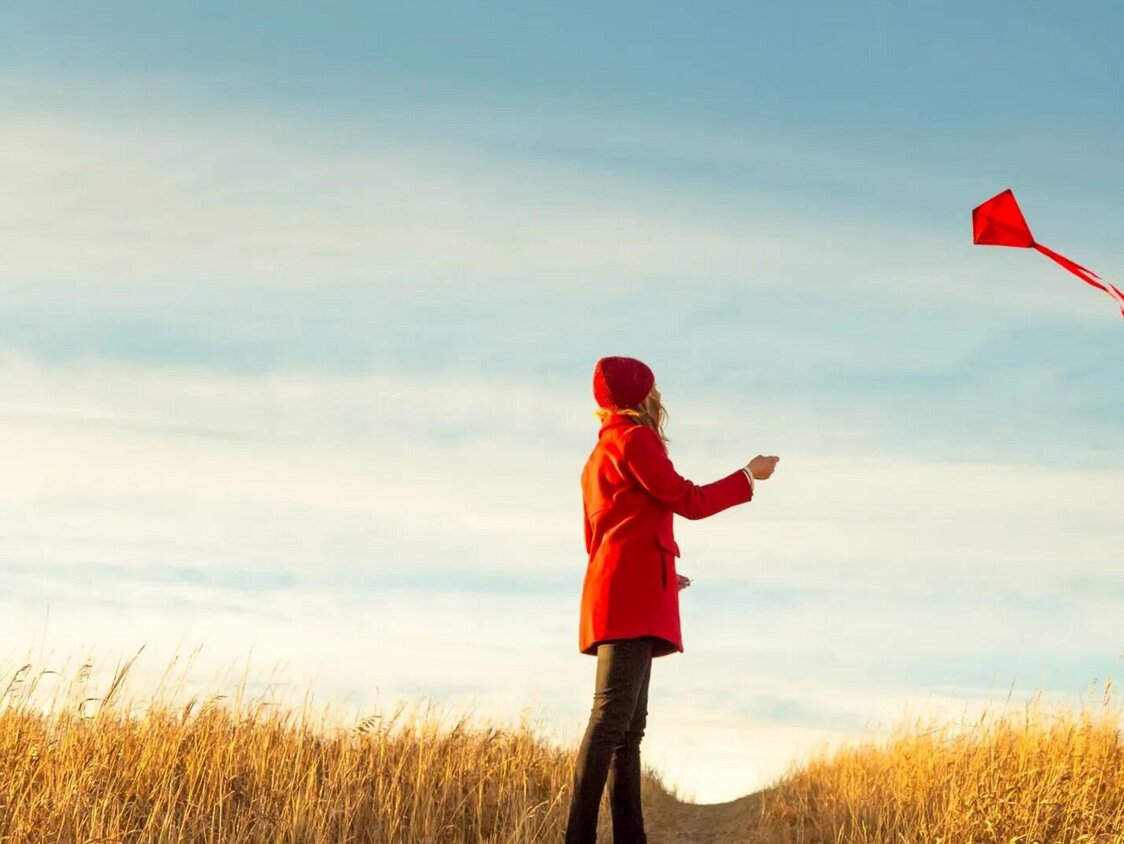 Eine Frau in einer roten Jacke lässt in einem herbstlichen Feld einen Drachen steigen.