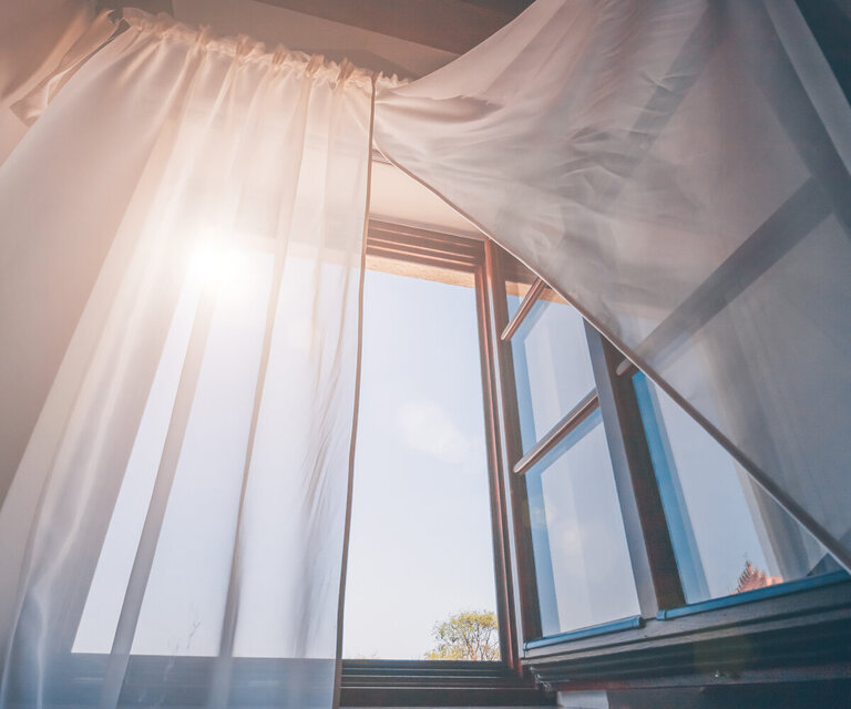  Ein geöffnetes Fenster, der Vorhang ist zur Seite gezogen und die Sonne scheint herein.