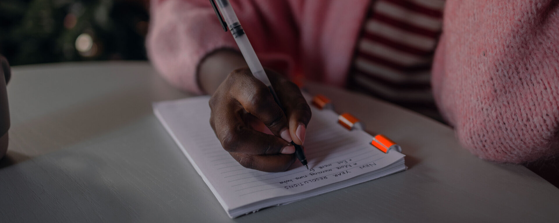 Eine Frau in einer rosa Strickjacke schreibt Neujahrsvorsätze auf einen Block.