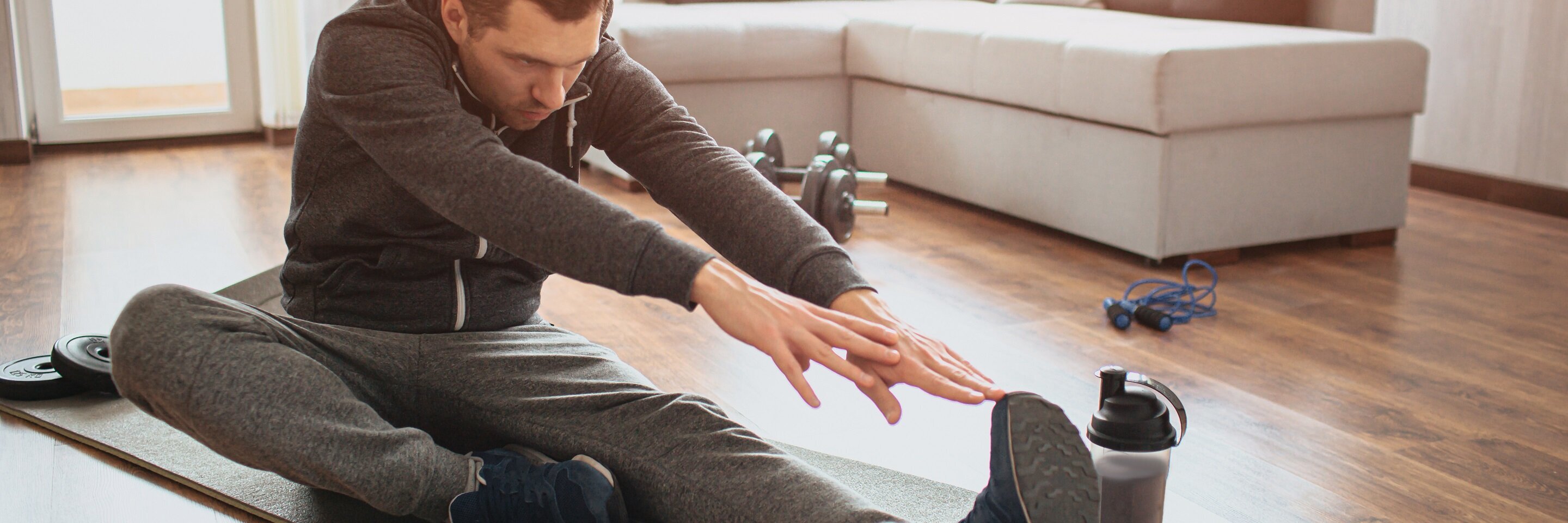  Ein Mann auf einer Matte im Wohnzimmer. Er sitzt und streckt sich, indem er mit beiden Armen Richtung ausgestreckter Fußspitze zieht. Im Hintergrund steht ein Sofa.