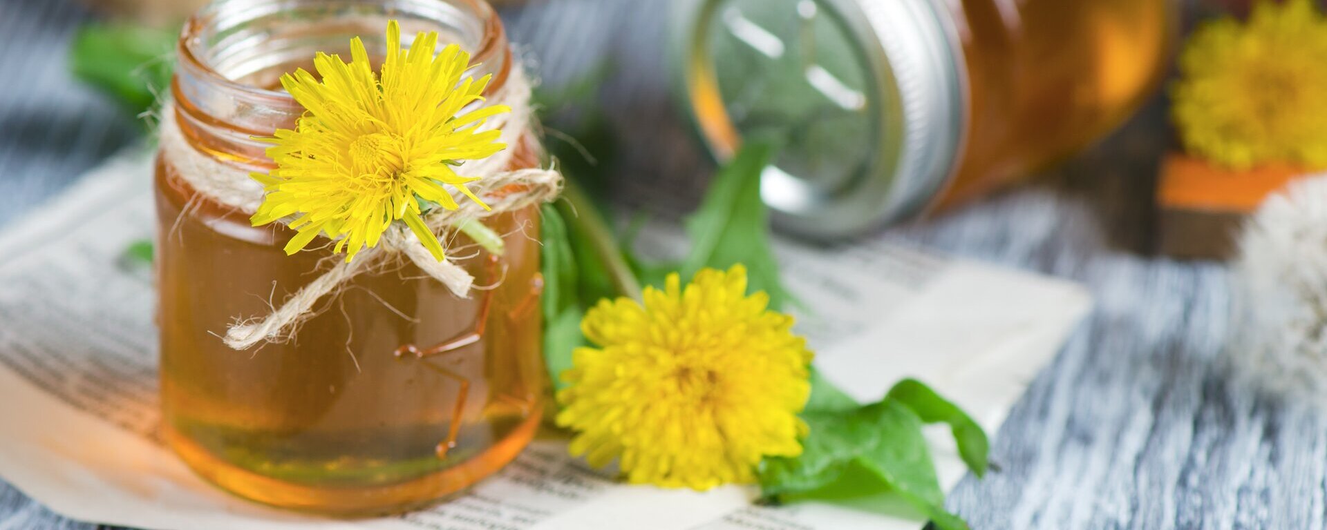 Zwei Gläser mit Honig und Löwenzahnblüten auf einem Tisch.
