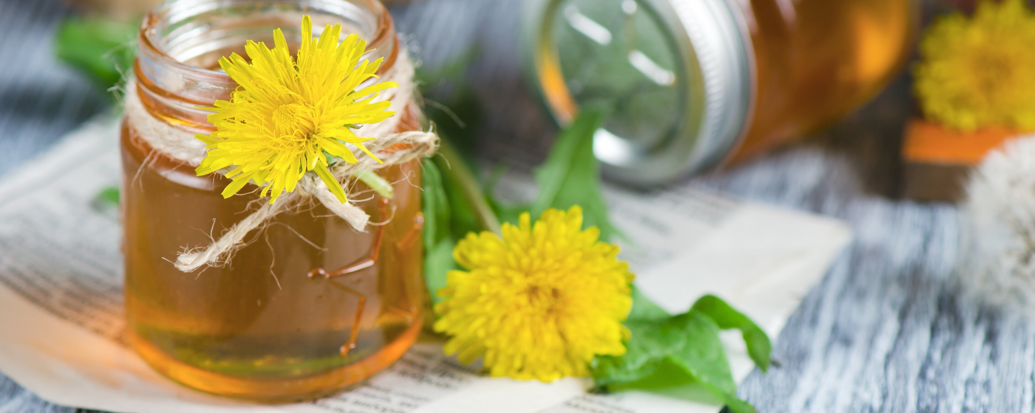 Zwei Gläser mit Honig und Löwenzahnblüten auf einem Tisch.