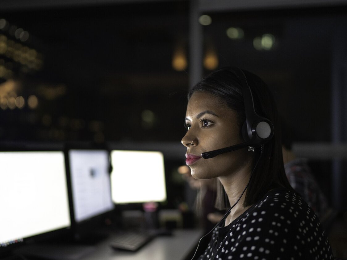 Eine Frau sitzt bei Dunkelheit vor einem Computer und trägt ein Headset.