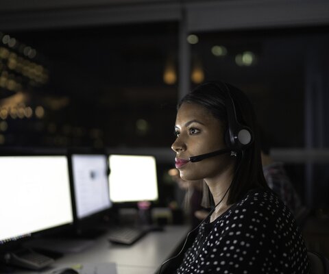 Eine Frau sitzt bei Dunkelheit vor einem Computer und trägt ein Headset.