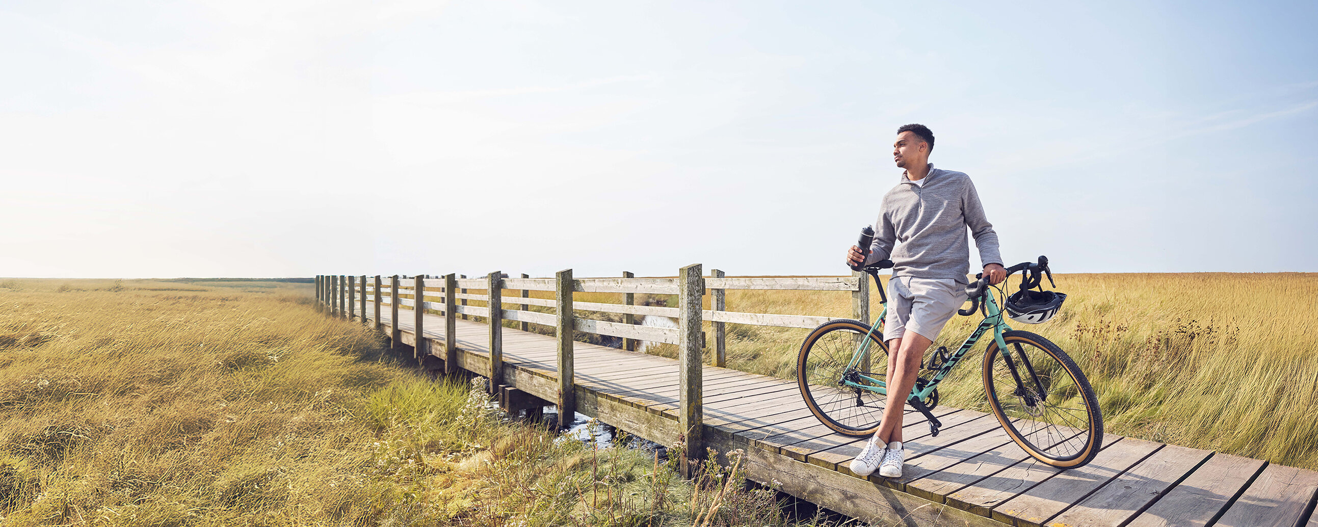 Ein Mann lehnt gegen sein Fahrrad auf einer Holzbrücke und hat eine Trinkflasche in der Hand.