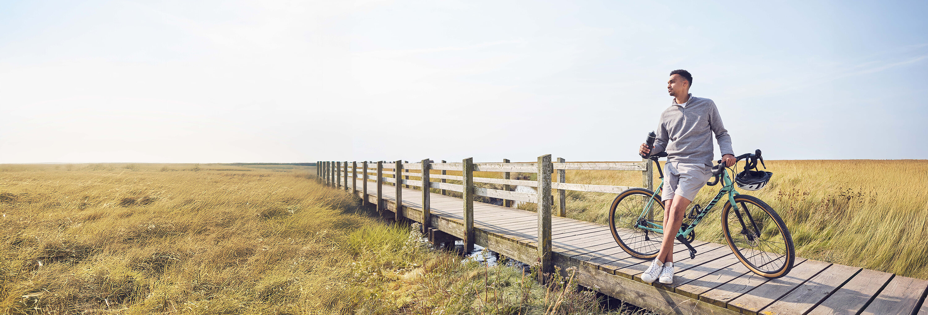 Ein Mann lehnt gegen sein Fahrrad auf einer Holzbrücke und hat eine Trinkflasche in der Hand.