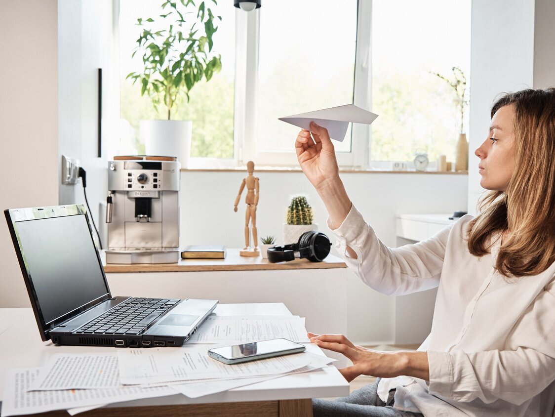 Eine Frau sitzt vor einem Laptop und hat einen Papierflieger in der Hand.