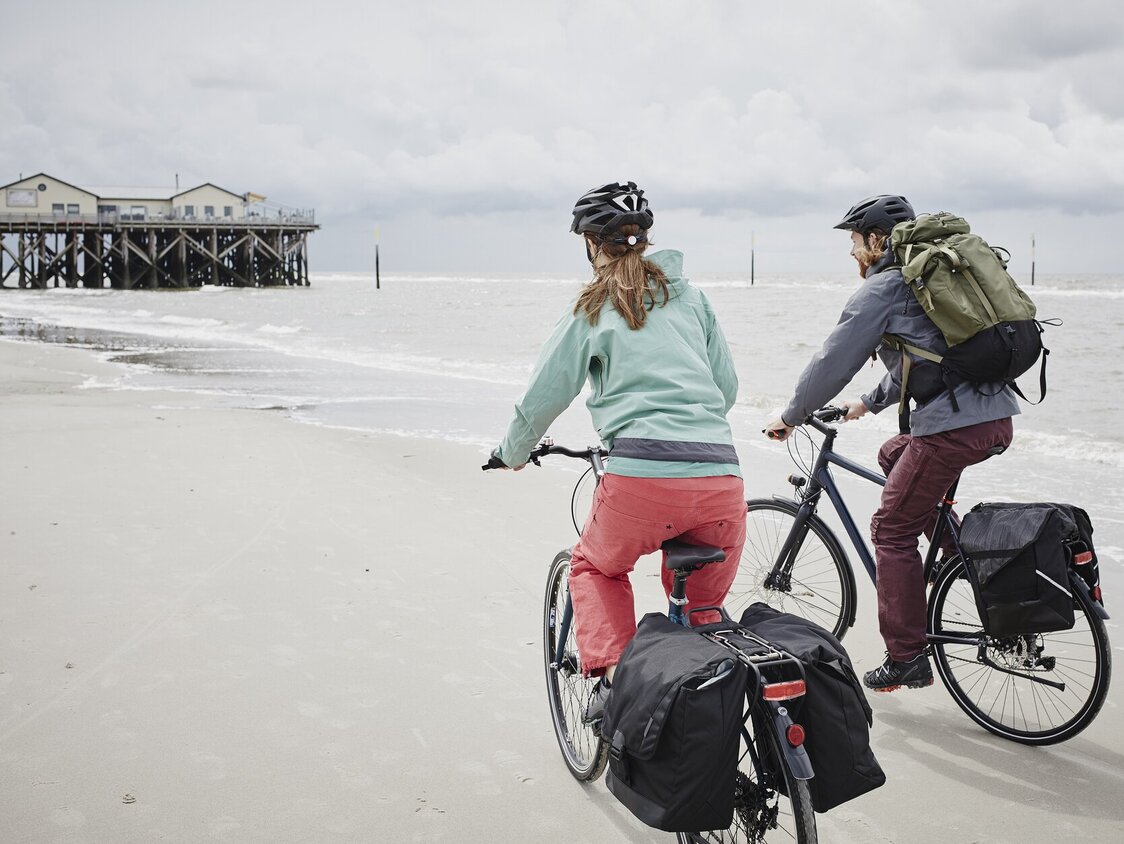 Ein Mann und eine Frauen fahren am Strand Fahrrad und haben viel Gepäck dabei.