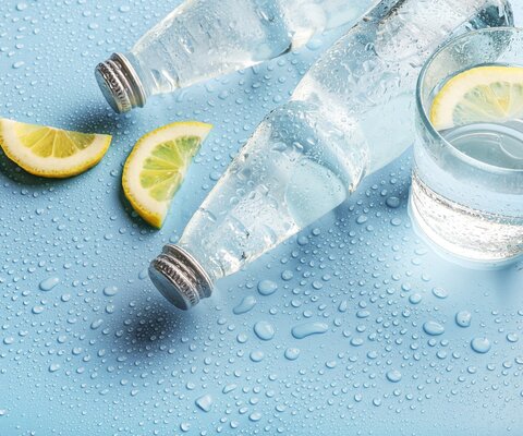 Zwei Flaschen Wasser und ein Glas mit Wasser auf blauem Untergrund. Daneben Zitronen.